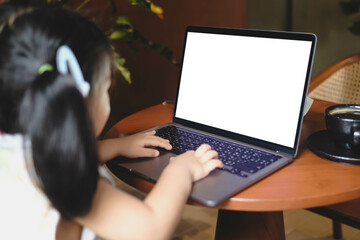 A young girl sits at a desk using a laptop with a blank white screen, ideal for showcasing...