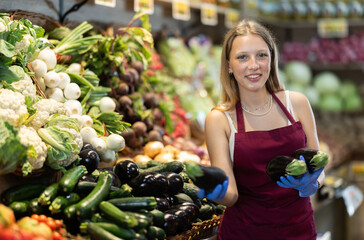 Shop seller put goods on display case. Girl creates pyramid of eggplant, presents fresh goods and...