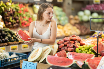 Woman in vegetable store choose half of watermelon. Client examine ripeness of fruit, take out fruits from showcase, collects grocery basket. Girl pull out ripest fruits from large pile of fruits.