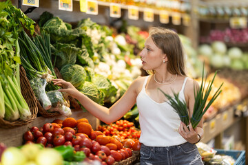 Girl client near vegetable stand in store choose bunch of green onion. Young woman shopping in...