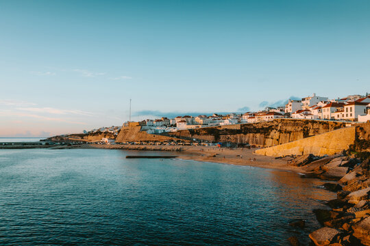 Golden hour sunlight illuminating the picturesque village of Ericeira atop cliffs overlooking the sandy beach and calm Atlantic waters in Mafra, Lisbon, Portugal