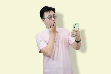 Young Asian man wearing glasses and a polo shirt, looking at his mobile phone with a confused and...
