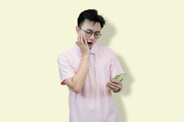 Young Asian man wearing glasses and a polo shirt, looking at his mobile phone with a confused and...