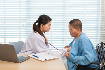 Young female doctor using stethoscope to examine senior woman in wheelchair during checkup, showing...