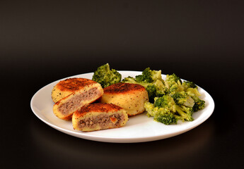 Potato cutlets stuffed with meat and boiled broccoli on a round ceramic plate on a black background.