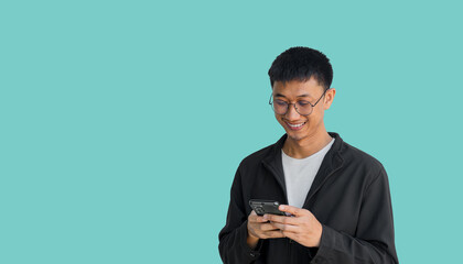 Young Asian man in his 20s wear glasses and wearing a black coat is happily playing on a mobile phone isolated on a blue background. Typing and chatting with friends via smartphone. Online social