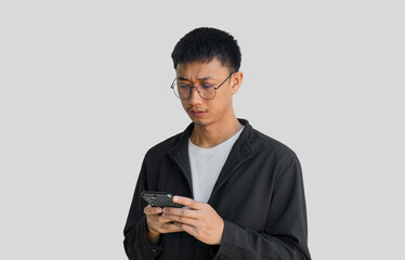 Young Asian man in his 20s wearing glasses and black coat looking at smartphone with serious expression on grey background. Social media stress concept. Negative energy. Social online.