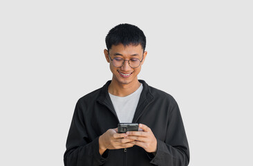 Young Asian man in his 20s wear glasses and wearing a black coat is happily playing on a mobile phone isolated on a grey background. Typing and chatting with friends via smartphone. Online social