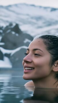 A smiling woman enjoying a geothermal spring. She is surrounded by water and snow capped rocks in the background. It is a serene and revitalizing scene of relaxation and nature's beauty.