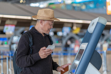 asian senior man passenger using smartphone,doing self check in at self service check in machine or KIOSK at the airport terminal,elderly retired with modern lifestyle,technology,transportation,travel