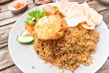 A plate of fried rice topped with over well fried egg, served with cucumber and tomato slices, lettuce, fried chicken and shrimp crackers
