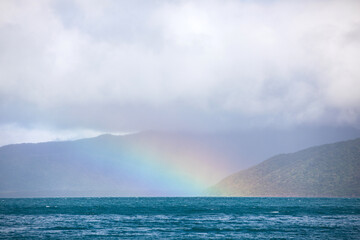 A faint rainbow arches from a cloudy sky onto a blue wind swept ocean adjacent a tropical coastal...