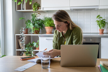 Focused woman reviewing bills, taxes, receipts on laptop with glass of water nearby. Financial...