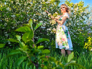Blonde woman posing in a floral dress near blooming flowers under clear blue skies