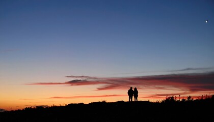 Silhouetted couple standing on a hill during sunset under a sky with colorful clouds and a crescent moon