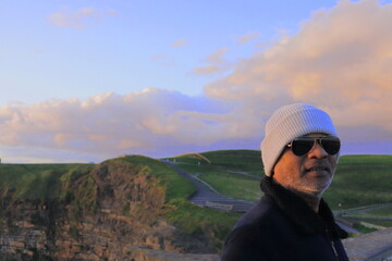 Naklejka premium Stylish Man at Cliffs of Moher on Bright Sunny Evening – Scenic View of Iconic Sea Cliffs and Atlantic Ocean in County Clare, Ireland with Clear Blue Sky and Dramatic Coastal Landscape