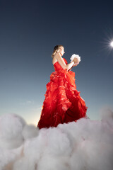A beautiful woman in a stunning red dress stands gracefully amid fluffy clouds against a vibrant,...