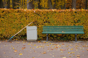 In the autumn park, there is a bench, next to an urn and a rake for cleaning leaves.