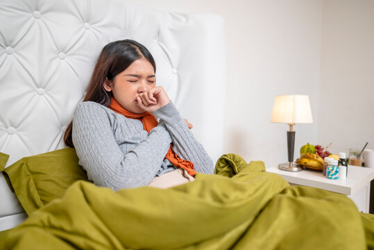 Indonesian southeast asian woman in a blanket sitting on the bed at home and coughing. Suffer from fever, flu, and cold. Healthcare concept