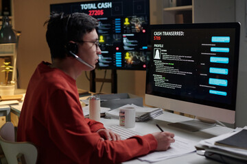 Young man wearing headset sitting at desk operating computer with scam call center interface on screen, engaging in fraudulent activity