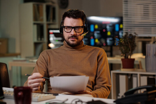 Portrait of Caucasian man wearing headset sitting at desk holding documents and pen looking into camera working in scam call center