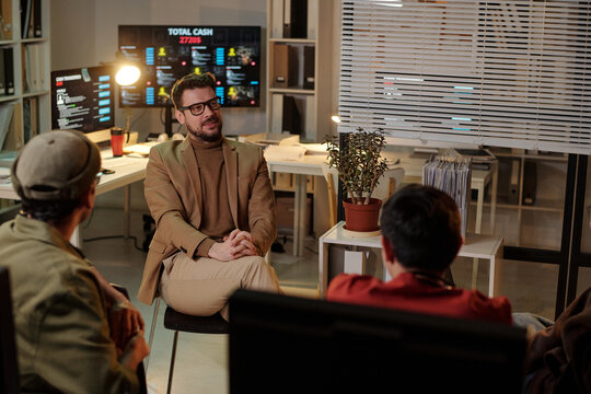 Caucasian man sitting and talking to two young adult men in office with computer monitors displaying financial data, scam call center context - Powered by Adobe