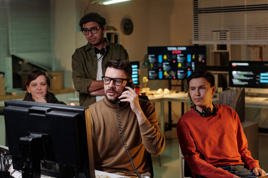 Group of multiethnic men and woman working in scam call center, one fraudster speaking on phone while others observing computer monitor