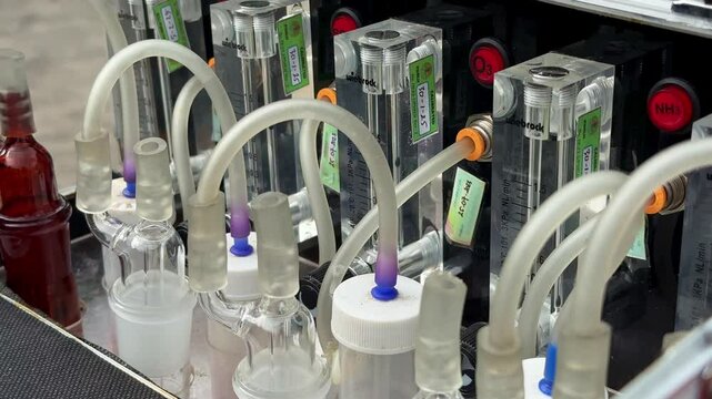 Detailed laboratory setup showing air sampling system with gas flow regulators for NO₂, SO₂, and O₃, flexible tubes, and reagent bottles for pollutant monitoring. Impinger machine.