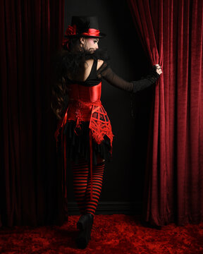 Burlesque circus performer, female model, in red corset and striped stockings in jester clown costume with top hat. Velvet stage curtains striking a theatrical full body pose gothic fantasy background
