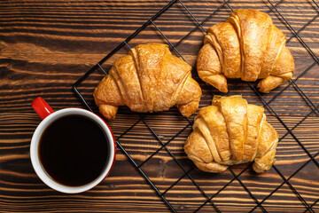 Freshly baked croissants on a cooling tray and a cup of coffee with a wooden background. Top view