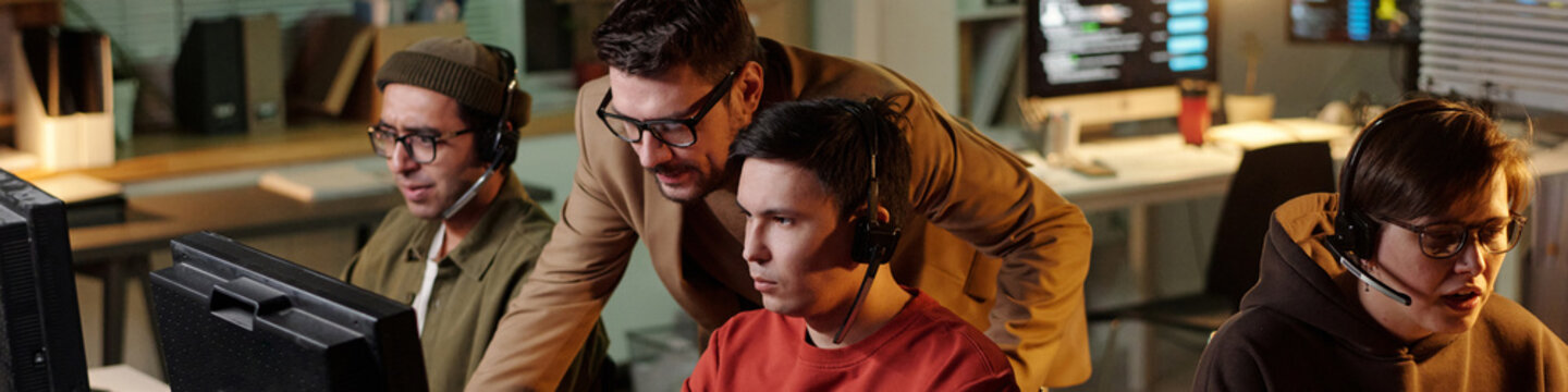 Multiethnic group wearing headsets operating computers in scam call center, Caucasian man supervising and instructing team during fraudulent activity