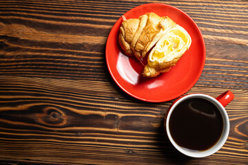 Slice of freshly baked croissant on a red plate with a cup of coffee on a wooden background. Top view