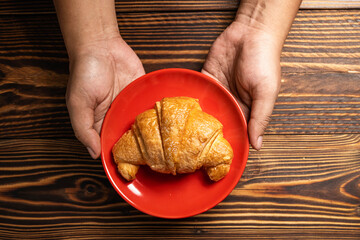 Human hand holding a freshly baked croissant on a red plate with a wooden background. Top view