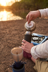 Person pouring hot water from a kettle into a thermos, outdoors, on the bank of a stream, at dawn....