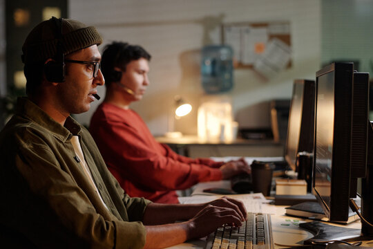 Young adult men wearing headsets operating computers, both sitting in dimly lit scam call center environment