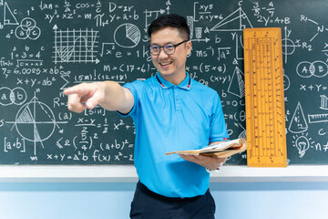 Asian math teacher smiling and pointing forward holding book in classroom with blackboard of...