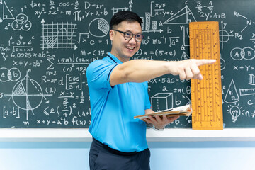 Asian math teacher smiling and pointing to side holding book standing in classroom with blackboard...