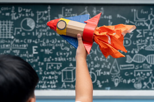 Student holding colorful handmade rocket in classroom with blackboard full of science and math formulas in background