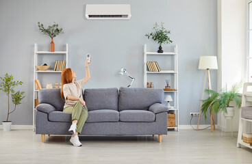 Portrait of a young smiling woman turning on modern air conditioner sitting on sofa at home....