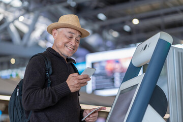 asian senior man passenger using smartphone,doing self check in at self service check in machine or KIOSK at the airport terminal,elderly retired with modern lifestyle,technology,transportation,travel