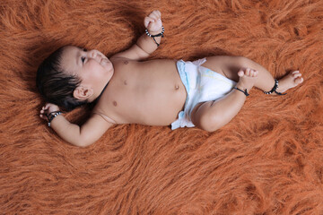 Cute Indian baby lying on a soft textured brown rug, wearing a diaper and traditional black thread...
