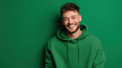 Fototapeta premium Cheerful young male with a casual green hoodie smiling broadly, standing before a vibrant green wall.