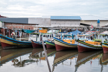 Traditional fishing village in Pangkalpinang with colorful wooden boats docked closely in calm waters, surrounded by modest waterfront buildings and lush greenery.