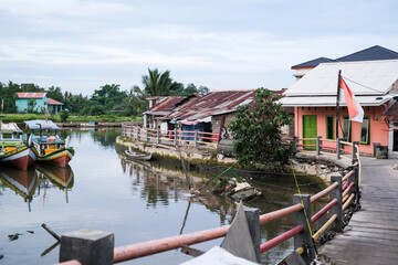 Traditional fishing village in Pangkalpinang with colorful wooden boats docked closely in calm waters, surrounded by modest waterfront buildings and lush greenery.
