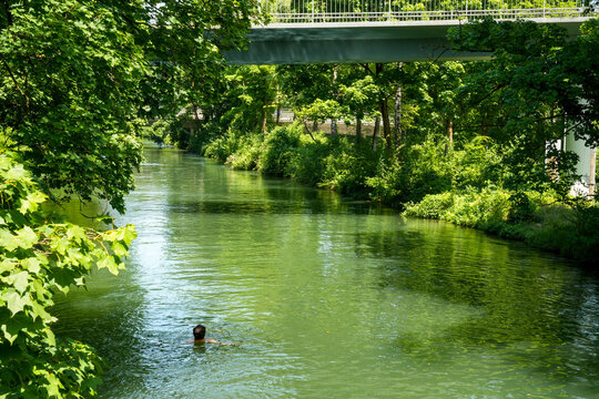 Floating man in the Eisbach (Isar), English Garden, Munich. Idyllic river landscape lined with lush greenery. A bridge spans the river. It is a tranquil and natural scene that invites you to linger