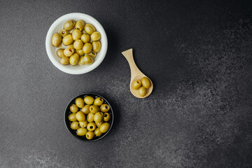 Two bowls with green olives and wooden spoon on dark textured background