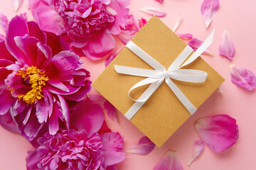 Gift box with white ribbon and pink peonies on pastel background with petals