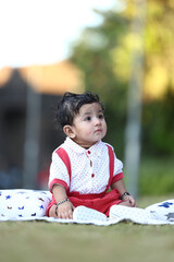 Adorable baby boy sitting on a grassy field with soft printed cushions, wearing a white and red...