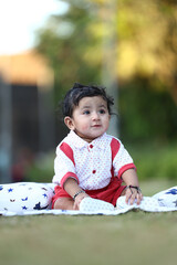 Adorable baby boy sitting on a grassy field with soft printed cushions, wearing a white and red...