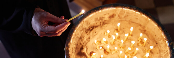 Header of male priest lighting candle in church, hand visible holding thin candle above large...